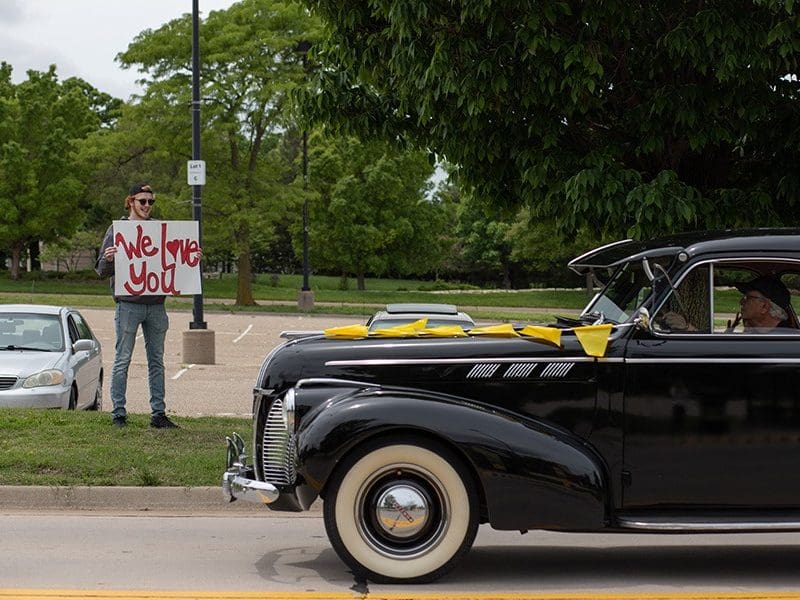 Dean Rodney Miller drives a classic car through campus, and a young man standing on the sidewalk holds a sign that says "We Love You."