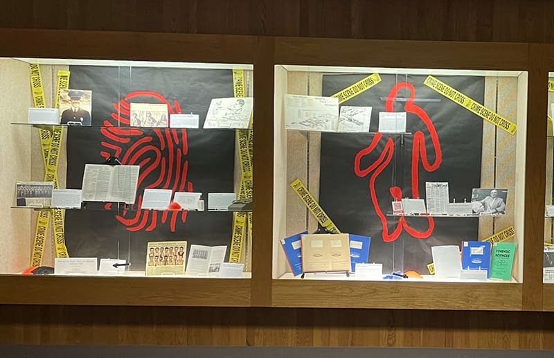 The images showes the exhibit "Crime Scene ICT" It is three different display cases with 2 glass shelves in each case with documents and photographs about the history of Forensic Science in Wichita. The backgound of each case is black with a large red "crime scene" related inmage in each. The first case on the black background is a red fingerprint. In the middle case on the black background is a red outline of a person. On the third case on the black background is a red microscope and vial. Each case has yellow crime scene tape that says "Crime Scene Do Not Cross."