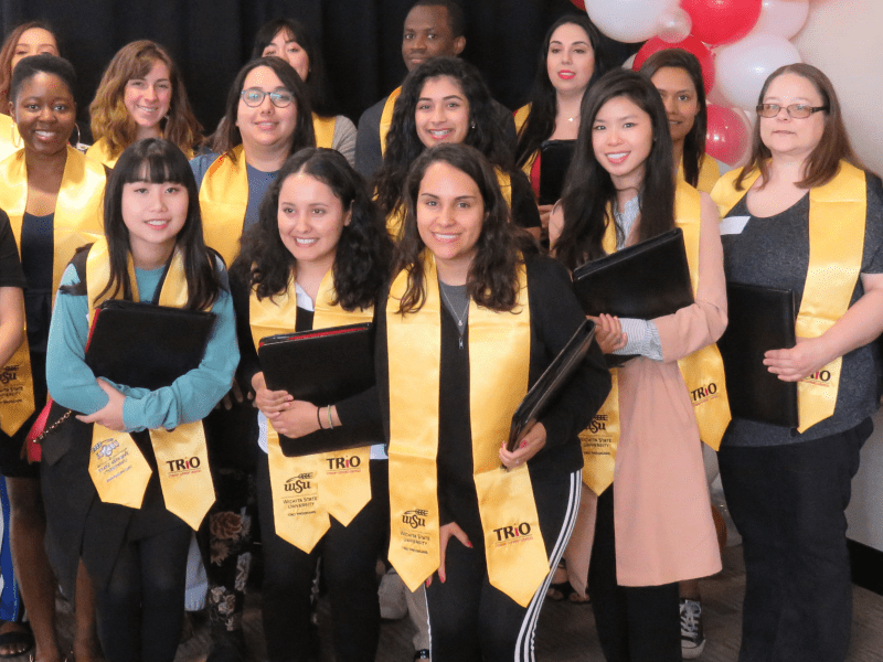 Several TRIO Student Support Services graduates gather for a group photo wearing their TRIO stoles and holding their graduate ceremony gift of a zippered pleather portfolio with big smiles.