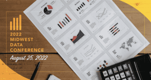 A wood table with a calculator and piece of paper featuring charts and graphics. 2022 Midwest Data Conference on August 25, 2022.
