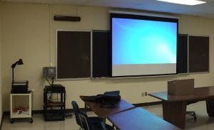 Photo of a classroom with a gray control box on the wall, as well as a transparency projector and projection screen.