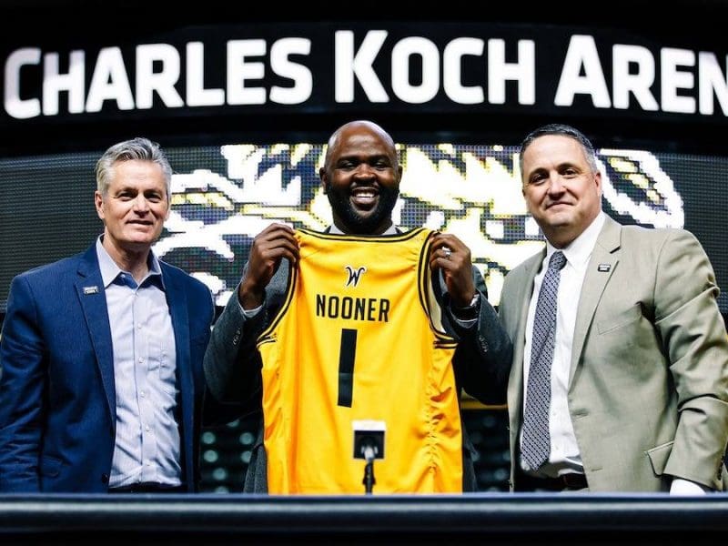 A photo of Terry Nooner holding up a Shocker jersey with President Rick Muma and Kevin Saal, director of athletics inside Charles Koch Arena.