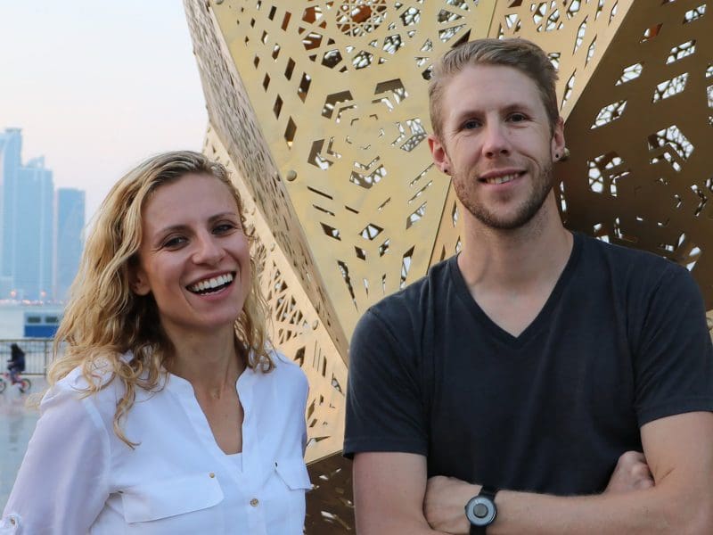 A photo of Yelena Filipchuk and Serge Beaulieu, from the art collective HYBYCOZO, standing in front of a many-sided, brass, geometric sculpture.