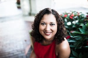A picture of a smiling Black woman with curly, shoulder length hair in a red dress in front of a background of flowers.
