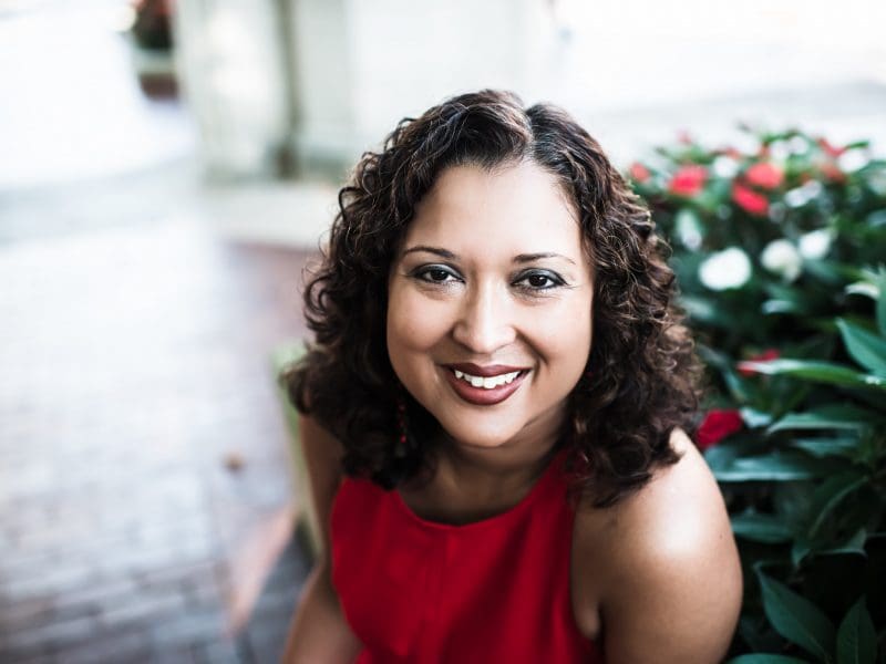 A picture of a smiling Black woman with curly, shoulder length hair in a red dress in front of a background of flowers.