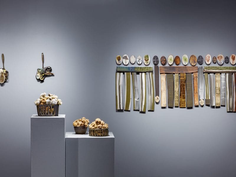 Ceramic works that look like baskets of cotton sit on two pedestals in front of six hanging ceramic works on a wall.