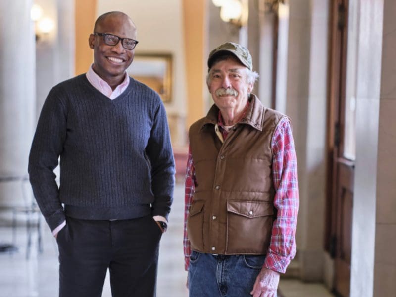 A middle-aged man and an older man pose indoors before some columns.