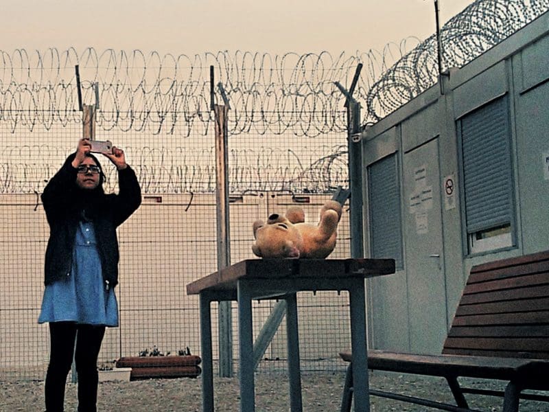 A girl in hijab takes a photo in an outdoor space surrounded by razor wire. A teddy bear sits nearby.