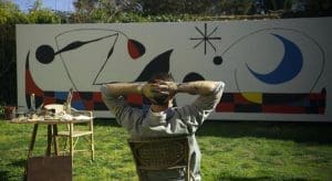 An artist sits with his back to us as he looks over a painting he is working on.