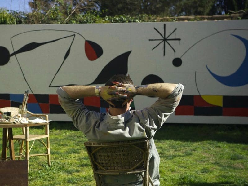 An artist sits with his back to us as he looks over a painting he is working on.
