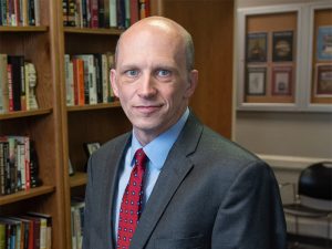 Dr. Andrew Orr from Kansas State University. Dr. Orr is a white man, wearing a suit and tie standing in front of a bookshelf.
