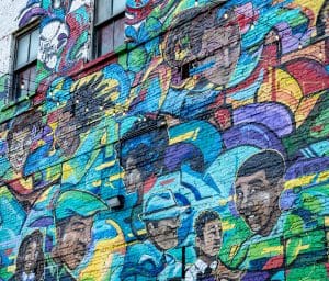 A mural by one of the Urban Canvas artists, Quintis Pinkston, is painted in spray paint and features smiling people.