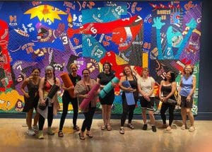 A group of yoga practitioners pose with their yoga mats in front of a large art quilt.