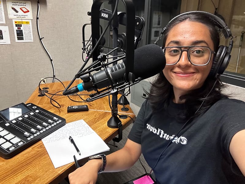 Image of Nidhi Shenoy behind a mic in a recording book with a notebook open in front of her. She's wearing a Sunflower t-shirt.