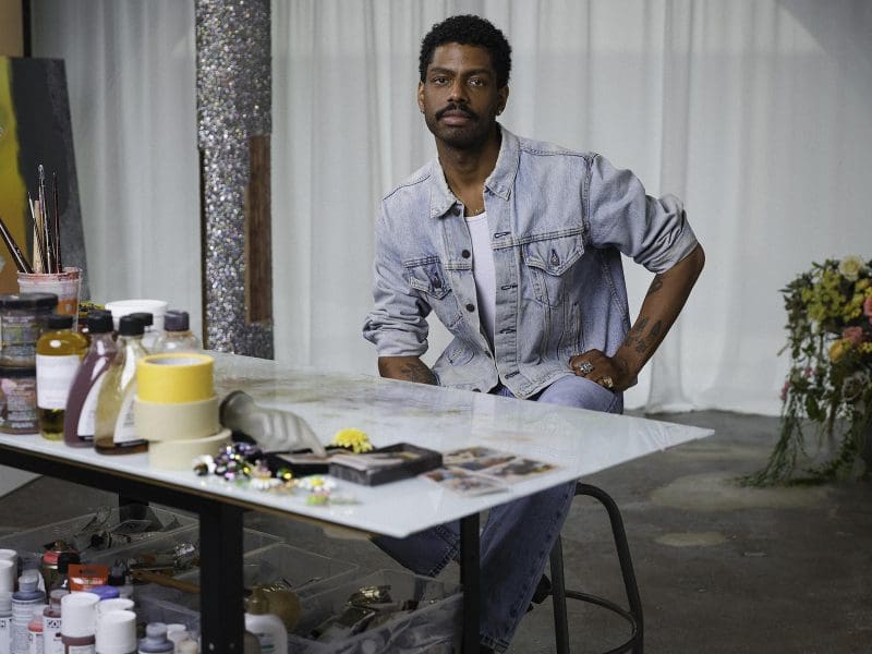 A young black, male-identifying artist is seen at a desk in his studio.