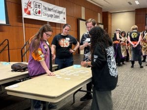 Two girls, one with long curly black hair wearing a black sweatshirt, and the second with long brown hair wearing a purple shirt with orange sleeves, stand around a table covered in cards with Japanese writing on them. Two judges stand behind them in black shirts with white name tags observing. In the back corner, a small group of five performers in costume can be seen standing in a circle talking. They wear long black tunics with gold and purple at the bottom and a yellow circle on the back.