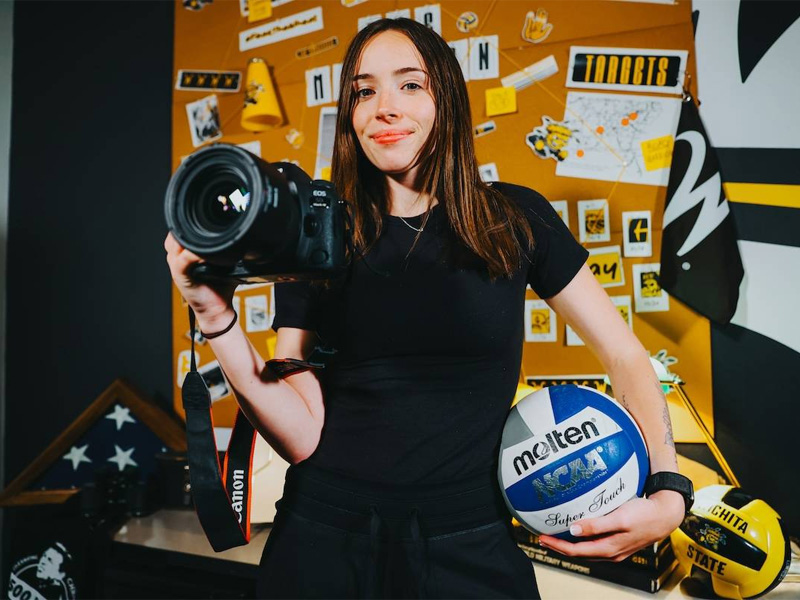 Skylar Fleeman, a WSU grad student who works in Shocker Athletics, poses with a camera and volleyball in front of a wall of Shocker Athletics gear