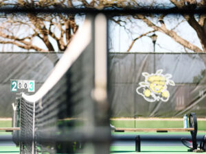 A profile shot of a tennis court net with benches and the WuShocker logo in the background. A board shows the score is 2 to 3.