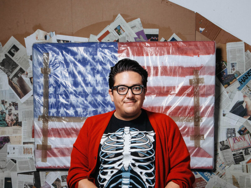 Artist Just Favela poses in front of an American Flag in a skeleton tee and red sweater