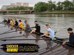 Alt Text: Shocker Rowing Camp.  A group of nine people are in a long, narrow rowing shell on a body of water, alongside a wooden dock. The people, who appear to be adults, are all holding oars and are dressed in casual athletic wear. They are seated in a line, facing the back of the boat, with only the person at the very front facing forward. Most of them are looking towards the camera, smiling. In the background, across the water, there's a bridge and a large, modern building. The sky is overcast.