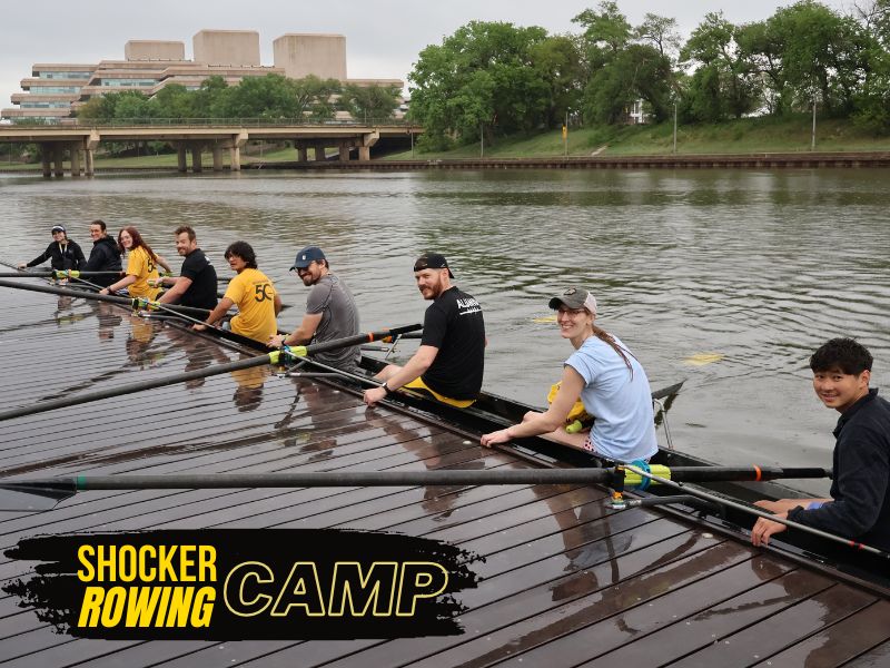 Alt Text: Shocker Rowing Camp.  A group of nine people are in a long, narrow rowing shell on a body of water, alongside a wooden dock. The people, who appear to be adults, are all holding oars and are dressed in casual athletic wear. They are seated in a line, facing the back of the boat, with only the person at the very front facing forward. Most of them are looking towards the camera, smiling. In the background, across the water, there's a bridge and a large, modern building. The sky is overcast.