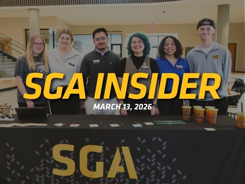 Six students stand behind a table with an SGA tablecloth inside a university building lobby. Large yellow text across the image reads “SGA Insider,” with “March 13, 2026” underneath. Items such as pens, cards, a laptop, and several iced drinks are arranged on the table, suggesting a student outreach or information table. The group smiles at the camera in a bright indoor campus space.