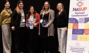 Stacy Chestnut, English teacher at East High School, Wichita Public Schools; Holly Ingram, assistant superintendent of learning services, Wichita Public Schools; Tammy Martin, executive coordinator of new teacher induction, Wichita Public Schools; and Heidi Stinchcomb, associate dean of the College of Education at Wichita State University, stand together on stage at the NASUP Annual Conference. Two members of the group hold blue glass award plaques, and a conference banner highlighting school-university partnerships is displayed beside them.