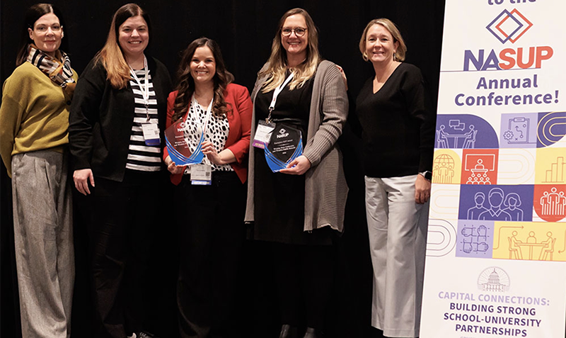 Stacy Chestnut, English teacher at East High School, Wichita Public Schools; Holly Ingram, assistant superintendent of learning services, Wichita Public Schools; Tammy Martin, executive coordinator of new teacher induction, Wichita Public Schools; and Heidi Stinchcomb, associate dean of the College of Education at Wichita State University, stand together on stage at the NASUP Annual Conference. Two members of the group hold blue glass award plaques, and a conference banner highlighting school-university partnerships is displayed beside them.