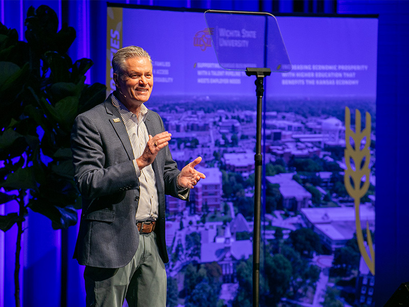 President Rick Muma stands on a stage while smiling mid clap. In the background are banners with photos of Wichita State University's campus and listing out the university's priorities.