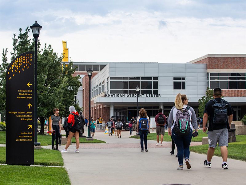 Students walking on campus, a sign with directions to various building sits off the path, and a building with the words Rhatigan Student Center on it is in the background.