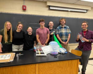 WSU science education students and a faculty member pose together behind a table of science demonstration materials at the KATS Kamp conference.