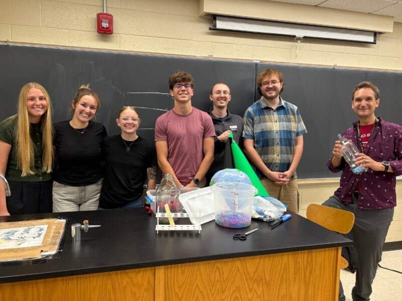 WSU science education students and a faculty member pose together behind a table of science demonstration materials at the KATS Kamp conference.