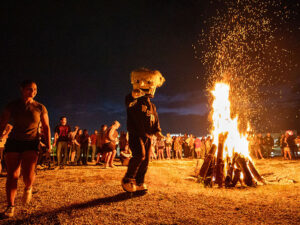 WuShock points to the camera standing next to a bonfire. Students are in the background.