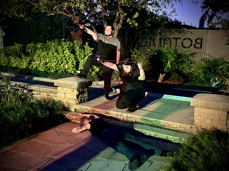 Three actors portraying a scene from Macbeth in Botanica's gardens. One actor holds a pop sword in the air. Another actor kneels on one knee with sword above their head. Third actor is face down on the ground.